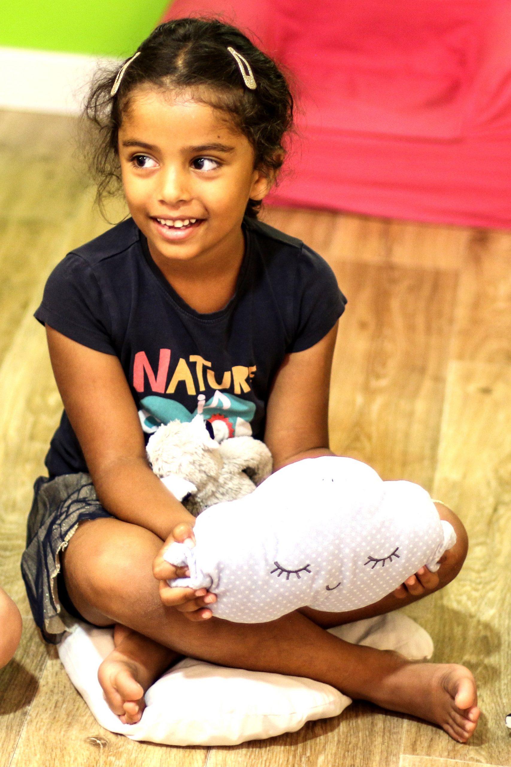 Cette image est une photo d'une fillette d'environ 8 ans, assise sur un coussin, tenant dans ses mains une peluche en forme de nuage.